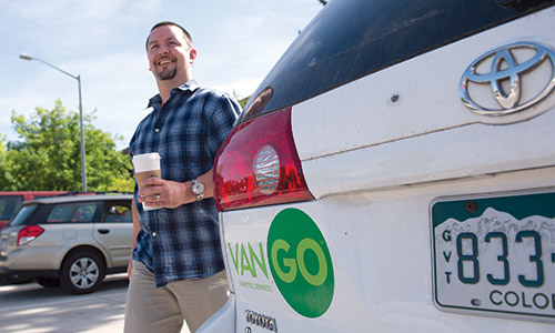 Photograph of a smiling man with a cup of coffee in his hand, standing next to a white vehicle with the VanGo logo.
