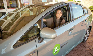 Photograph of a student in a gray Zipcar vehicle. She had her window, is looking out the window and smiling.