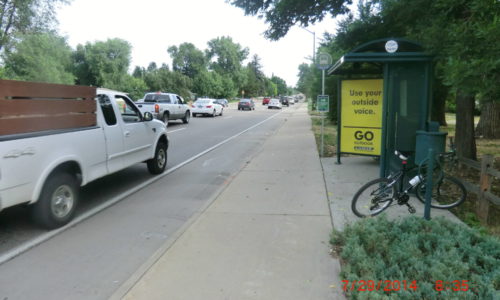 Photograph of a shelter TransFort bus stop next to a bust road with a bike rack next to the bus stop.
