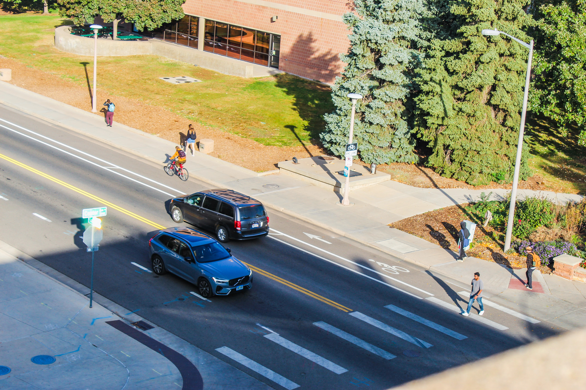 A street view on a sunny day near a university campus. Pedestrians walk along the sidewalk and cross at a marked crosswalk. A cyclist rides in a designated bike lane, and two vehicles drive down the road. Trees, a brick building, and a lamp post with a bicycle sign are visible in the background.