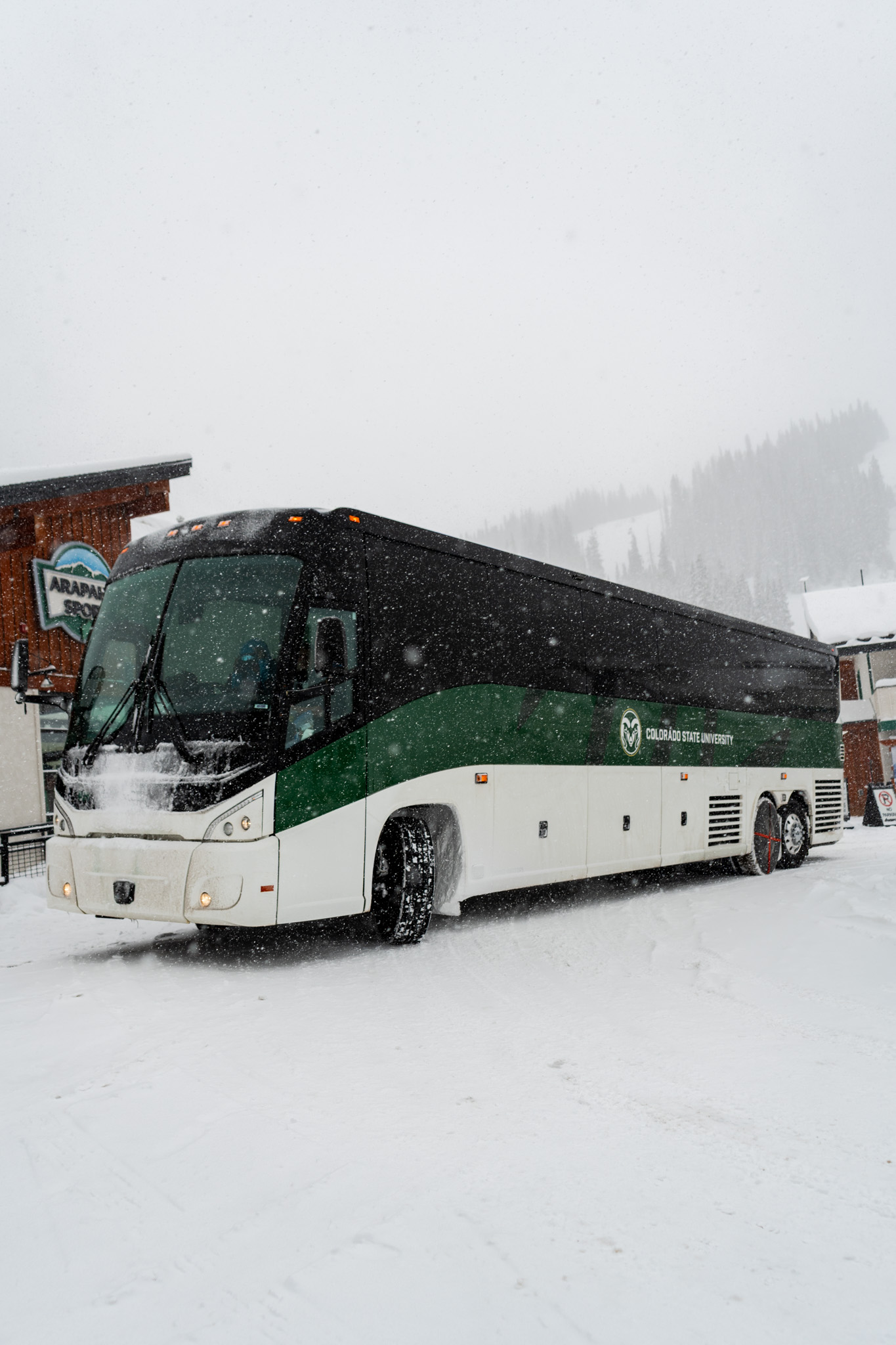A CSU green-and-white SKISU bus pulls into a snowy Arapahoe Basin area during heavy snowfall.
