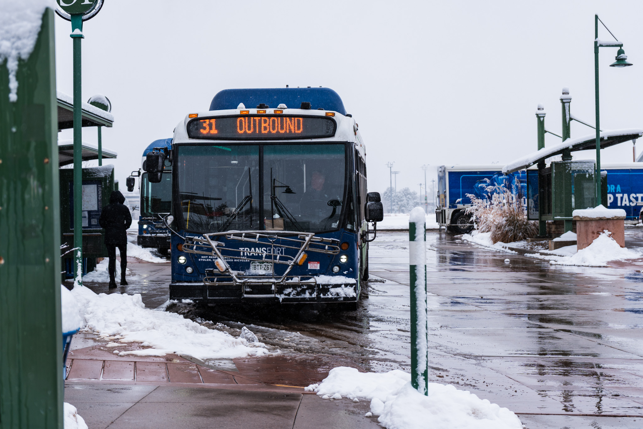 A Transfort bus displaying “31 Outbound” pulls into a snowy transit station, with slushy pavement, a person waiting at the stop, and snow-covered shelters and light posts surrounding the area.